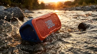 An orange JBL speaker splashing in a river at sunset, demonstrating the durability of rugged waterproof speakers.